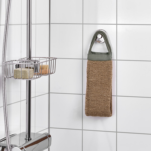Un exfoliante corporal de yute cuelga de un gancho en un baño, con asas de fácil agarre. Cerca de allí, un carrito de ducha cromado guarda artículos.