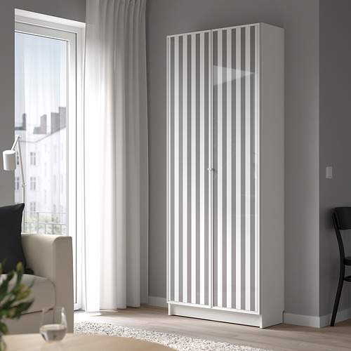 La estantería alta BILLY blanca con puertas de vidrio se eleva junto a la ventana. Diseño moderno y minimalista.