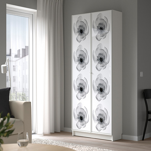 Sala de estar moderna con estantería alta blanca BILLY, adornada con diseños florales negros, de pie cerca de una gran ventana con cortinas transparentes.
