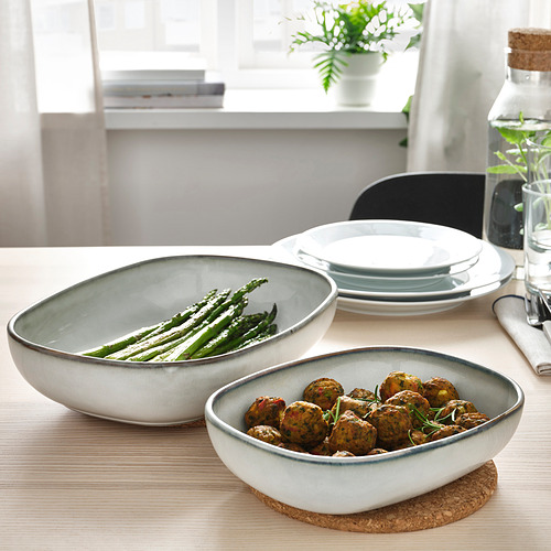 Dos cuencos aptos para horno con comida en una mesa. Uno tiene espárragos, el otro albóndigas. Forma parte de una serie GLADELIG elegante y duradera.