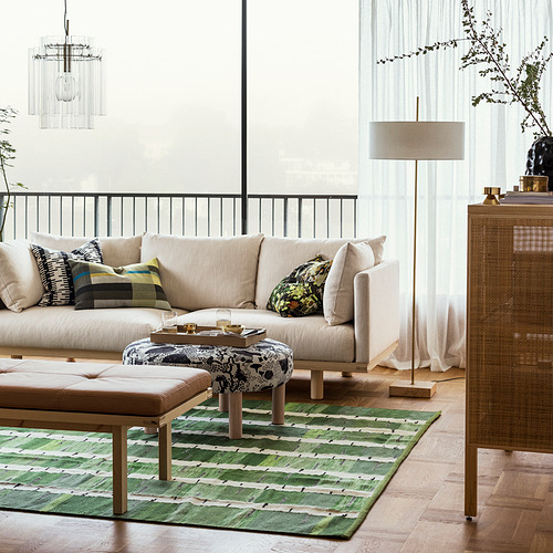 Moderno salón con sofá beige, alfombra verde estampada, elegante lámpara de pie con pantalla textil y base de latón junto a un gran ventanal.