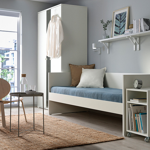 Dormitorio minimalista con sofá cama VIHALS blanco, cojines azules, muebles de almacenaje a juego, silla de madera y gran ventana.
