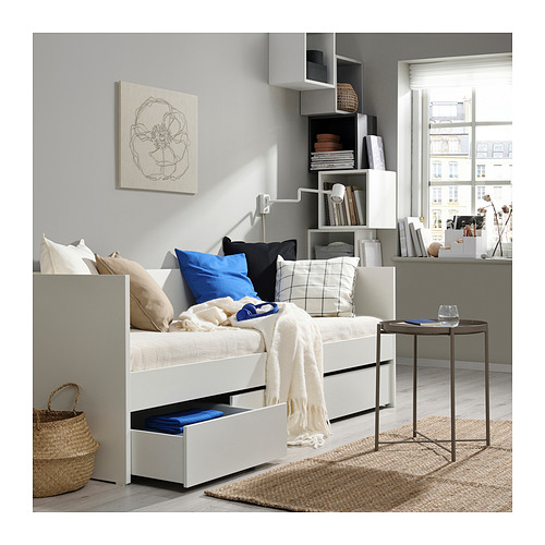 Dormitorio minimalista con sofá cama blanco y cajones. Almohadas de colores, alfombra y arte en la pared. Escritorio con silla, y vista a la ciudad desde la ventana.