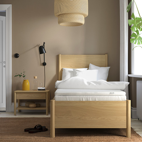 Dormitorio con paredes marrones, cama de madera enmarcada en roble claro con almohadas blancas y un jarrón amarillo en mesita de noche.