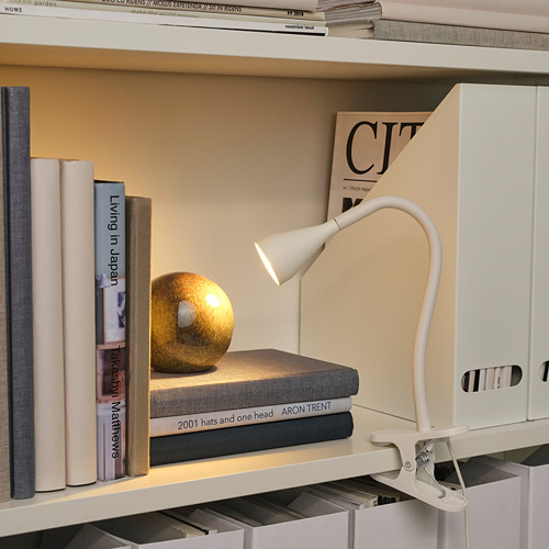 Estantería con libros y revistas, iluminada por una lámpara blanca de abrazadera ajustable. Un pequeño globo terráqueo descansa sobre los libros.