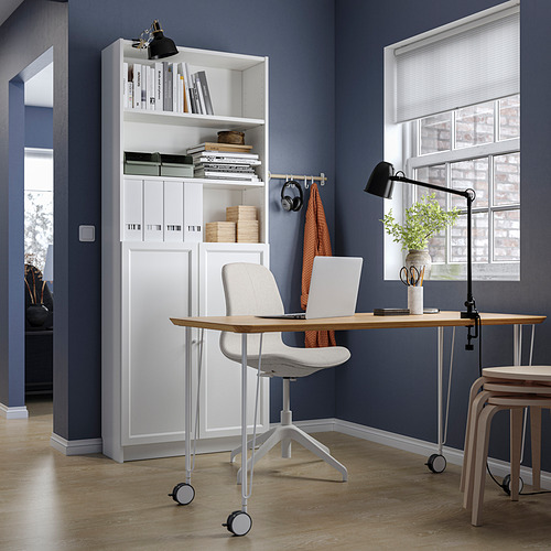 Moderna oficina en casa con una estantería blanca BILLY llena de libros y decoración, un escritorio de madera, una silla gris y una ventana con persianas.