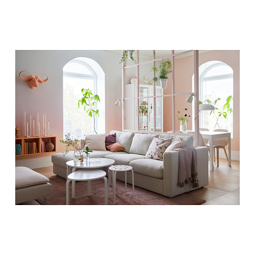 Moderno salón con sofá VIMLE en gris claro, cojines decorativos, mesas de centro blancas, grandes ventanales, plantas y una pared rosácea.