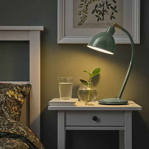 Acogedor dormitorio con lámpara de escritorio RÖDFLIK verde sobre una pequeña mesa al lado de la cama, con pantalla ajustable, vaso de agua y un pequeño jarrón de plantas.