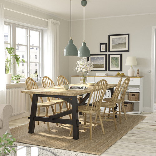 Comedor moderno con mesa de madera, sillas y grandes lámparas colgantes verdes. La decoración incluye fotos enmarcadas y un jarrón de flores alto. Cortinas blancas.