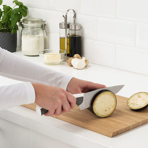 Persona cortando berenjenas en una tabla de cortar con cuchillos de cocina, rodeada de recipientes y una planta.