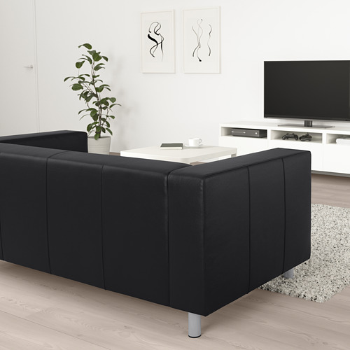Sofá KLIPPAN símil cuero negro, salón moderno y minimalista. Televisor de pantalla plana a la derecha, pared blanca, planta al lado del sofá.