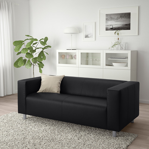 Sala de estar moderna con sofá KLIPPAN negro, almohada beige, gabinete blanco y planta grande.