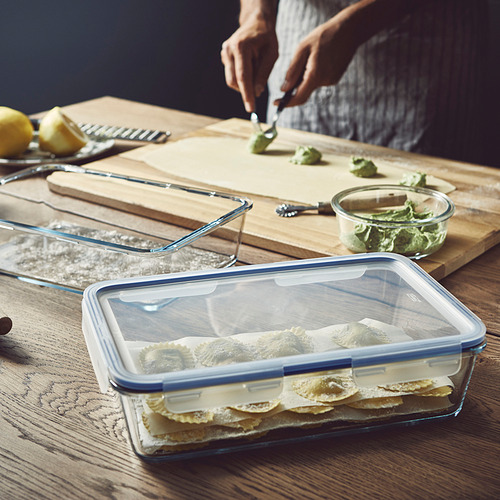 Persona que prepara la comida en una tabla de madera. Un recipiente de vidrio con tapa está lleno de platos en capas, probablemente sobras, listos para almacenar o transportar.