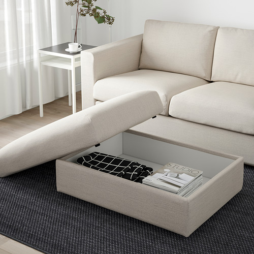 Sofá modular VIMLE beige con otomana de almacenamiento abierto, lleno de revistas debajo de una alfombra gris.