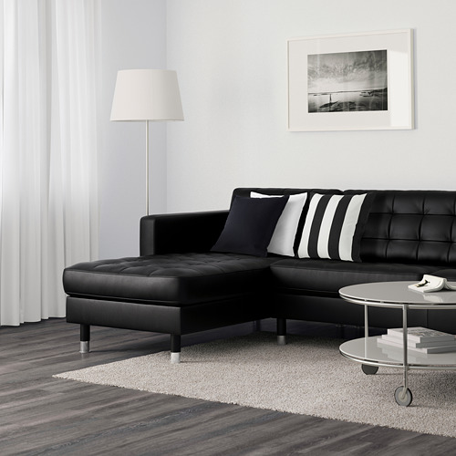 Sala de estar moderna con sofá seccional de cuero negro, almohadas a rayas, lámpara de pie, mesa de café con libros y obras de arte enmarcadas en la pared blanca.