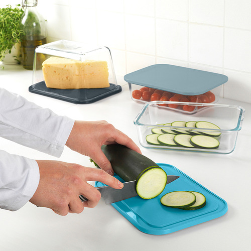 La persona corta el calabacín en una tabla de cortar/tapa azul. Cerca de allí, queso y tomates almacenados en contenedores IKEA 365+.