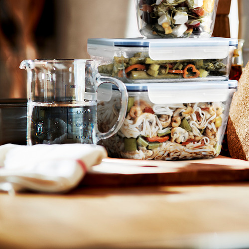Recipientes de vidrio apilados con tapas con cerradura, uno con verduras y fideos, colocados sobre una mesa de madera junto a una jarra medidora de agua.