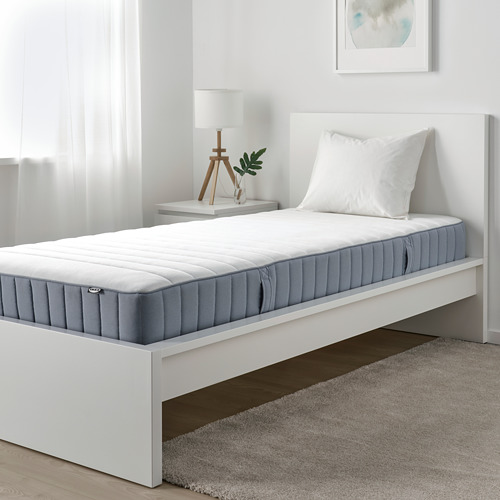 Dormitorio minimalista con colchón VALEVÅG de IKEA, somier azul grisáceo, blanco y lámpara de planta pequeña.