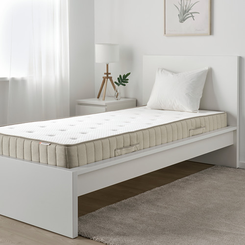 Cama individual minimalista blanca con colchón vatnestrom y almohada blanca. Mesita de noche con lámpara a un lado. Alfombra sobre suelo de madera.