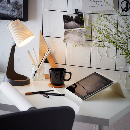Espacio de trabajo moderno con escritorio blanco, taza negra, tableta, lámpara, bolígrafos y planta.