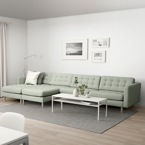 Moderno salón con gran sofá LANDSKRONA verde en forma de L, diseño capitoné, mesa de centro blanca, alfombra, fotos en blanco y negro y lámpara de pie de esquina.