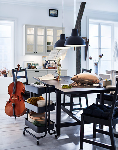 Comedor luminoso con sillas negras NORDVIKEN, mesa de madera, luces colgantes, instrumentos musicales y cocina al fondo.