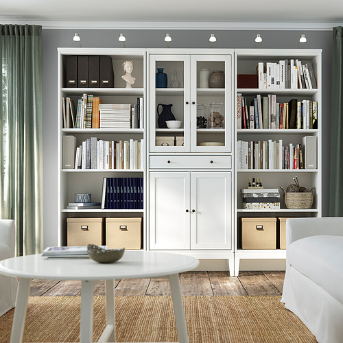 Estanterías y armarios blancos de IDANÄS llenos de libros y artículos decorativos, sobre una pared gris con cortinas verdes.