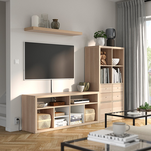 Una sala de estar moderna con un mueble KALLAX de madera clara con estantes abiertos, cajones y un televisor montado en la pared.