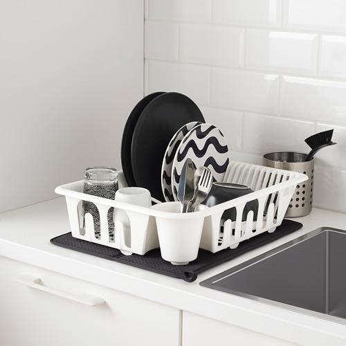 El estante de platos blanco en la encimera de la cocina contiene tazas, sartenes y utensilios. La alfombrilla de secado negra debajo atrapa las gotas.