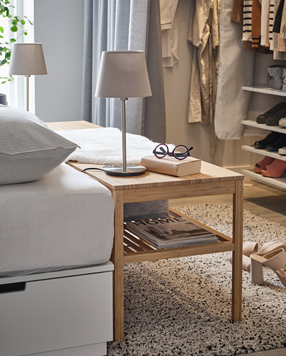 Lámpara de noche sobre mesa de madera cerca de la cama, con revistas y gafas.