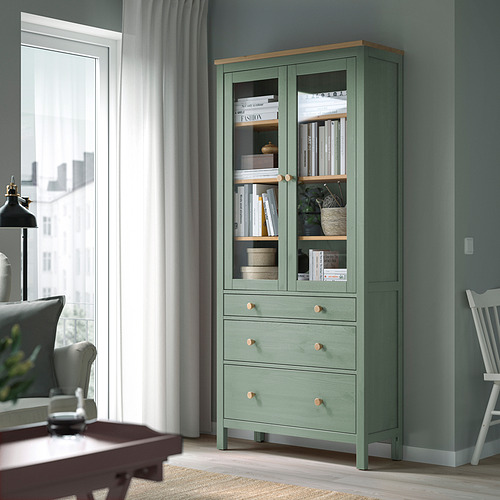 La estantería verde HEMNES con puertas de vidrio se alza cerca de la ventana, rodeada de sillas y cortinas, mostrando libros y decoración.