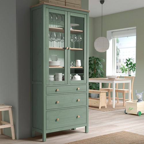 Mueble verde HEMNES con puertas de cristal, almacenamiento de platos y vasos en un comedor de madera clara.