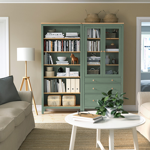 Acogedora sala de estar con una gran estantería HEMNES verde, lámpara de pie de madera, alfombra beige y sofá crema. Libros y artículos de decoración dispuestos ordenadamente en los estantes.