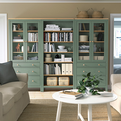 Librería HEMNES verde grisácea con libros y decoración entre sofás beige y mesa blanca