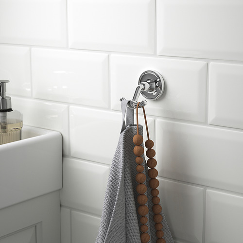 Un doble gancho cromado en la pared de un baño con azulejos sostiene una toalla gris y un collar de cuentas de madera, cerca de un lavabo blanco con dispensador de jabón.