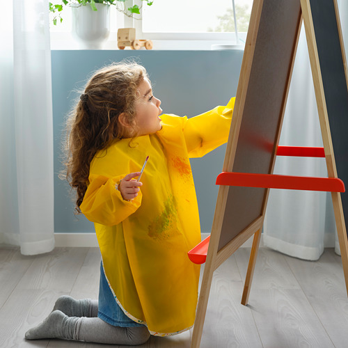Niña con un delantal amarillo de MÅLA, pintando en un caballete con marco rojo, mostrando la función protectora de los delantales.