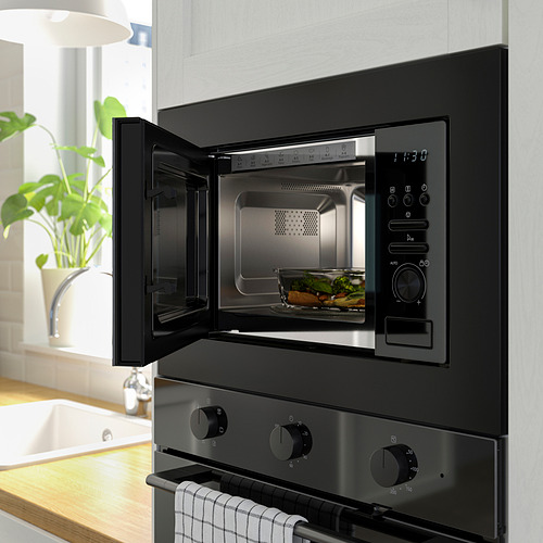 Escena de cocina moderna: microondas empotrado negro abierto con comida adentro, encima de la estufa negra con perillas, al lado de la planta en maceta.