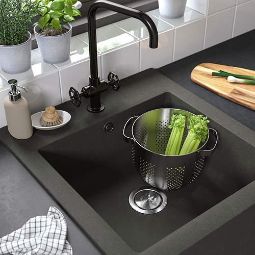 Fregadero de granja negro con cesta coladora de acero inoxidable rellena de apio. Cocina moderna con grifería negra y varios accesorios.