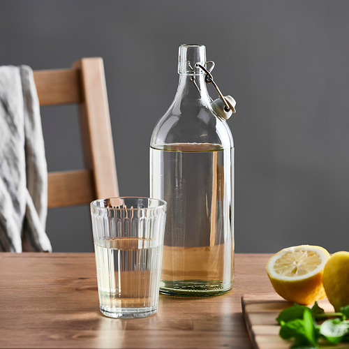 Botella de vidrio transparente con tapón en mesa de madera junto a vaso lleno y limones.