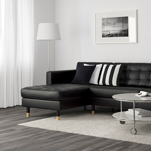 Sala de estar moderna con sofá en L negro, almohadas a rayas / sólidas, lámpara blanca, mesa de libros, alfombra grande sobre piso de madera oscura.