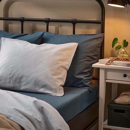 Acogedora cama con almohadas DVALA de color azul grisáceo y detalles a rayas blancas, con una cabecera de metal negro y una pequeña mesita de noche al lado.