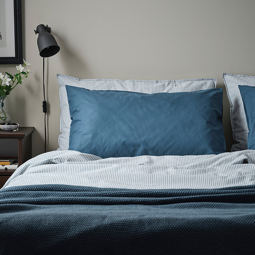 Una cama con almohadas azules DVALA. Las almohadas son rectangulares, de algodón suave, y se ajustan perfectamente a la cama, contrastando con un diseño de ropa de cama a rayas.