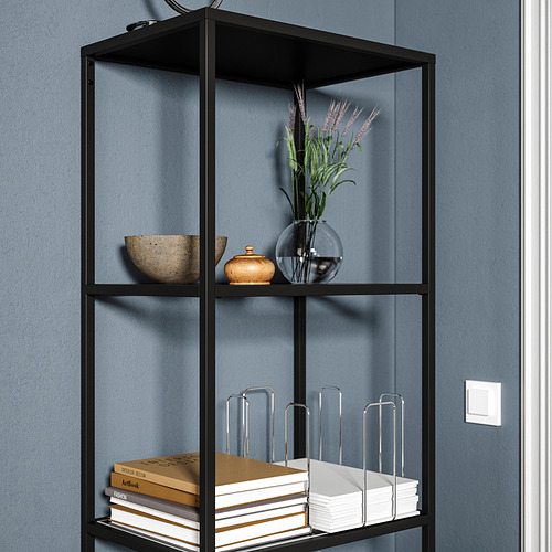 Estante de metal negro con vidrio, que sostiene libros, cuenco decorativo, jarrón y flores contra la pared azul.
