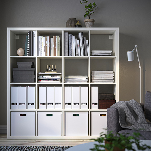 Estantería KALLAX con libros, revistas y cajas junto a un sofá gris.