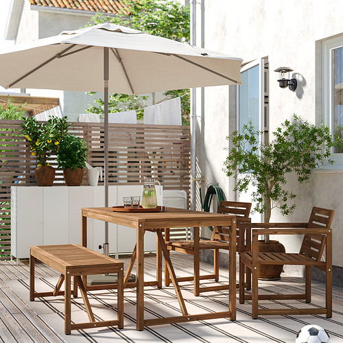 Patio exterior con mesa de madera NÄMMARÖ, sillas, banco y sombrilla beige. Rodeado de plantas y un edificio blanco.