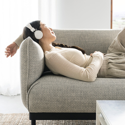 Mujer relajante en un sofá gris ÄPPLARYD, con auriculares, manta acogedora, decoración neutra.