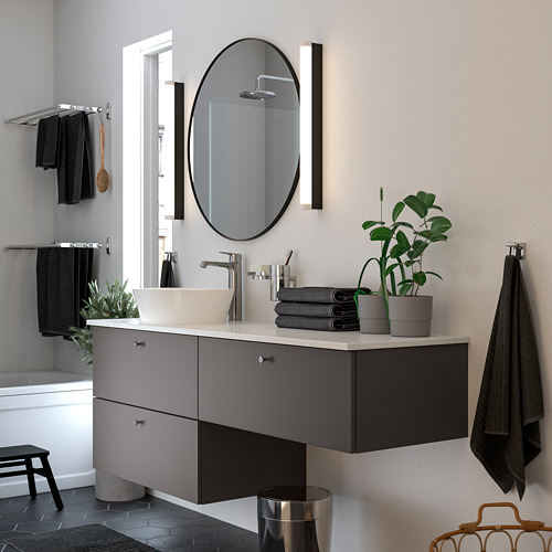 Baño moderno con espejo LINDBYN, encimera gris, lavabo blanco, accesorios de plata, planta verde y toallas negras.