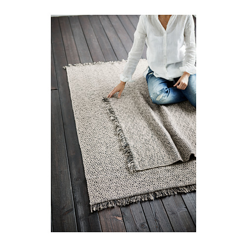 Persona arreglando una alfombra de algodón RÖRKÄR beige y negro sobre suelo de madera oscura.