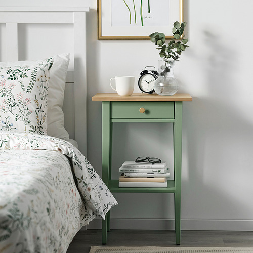 Mesita de noche verde con ropa de cama floral, taza, reloj, jarrón, libros y vasos. Impresión de la planta arriba.
