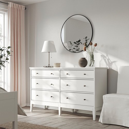 Cómoda minimalista IDANÄS con 6 cajones, lámpara y jarrón. El espejo refleja la decoración de la habitación. Pisos y cortinas de madera.